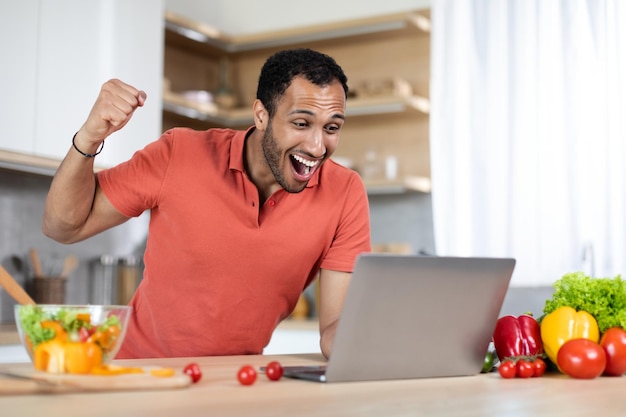 Un afro-américain du millénaire excité en riant à table avec des légumes biologiques utilise un ordinateur portable et fait un geste de réussite