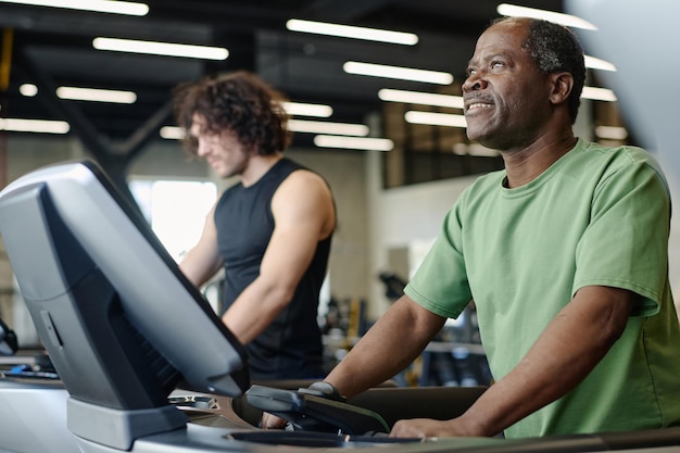 Un Afro-Américain marche sur un tapis roulant au gymnase avec un sportif caucasien en arrière-plan