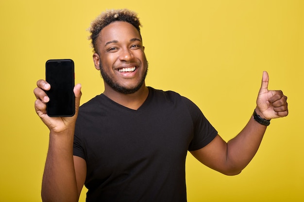 Un afro-américain musclé montre l'écran d'un téléphone moderne avec une place pour une mise en page Un homme noir satisfait sourit et donne un coup de pouce sur un fond jaune