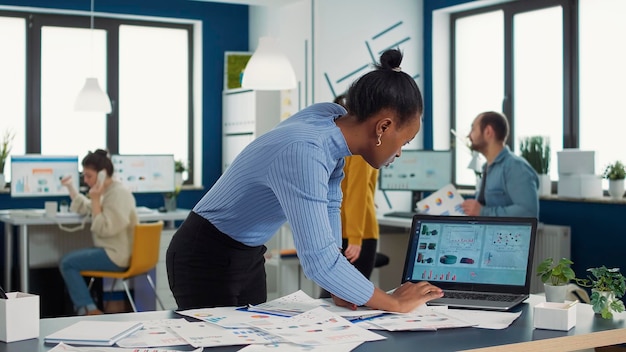 Afro-américain occupé en démarrage mettant le presse-papiers sur le bureau en regardant des papiers avec des graphiques de vente comparant les résultats avec les données sur l'écran d'ordinateur portable. Analyste d'affaires à la recherche de statistiques lors de la saisie.