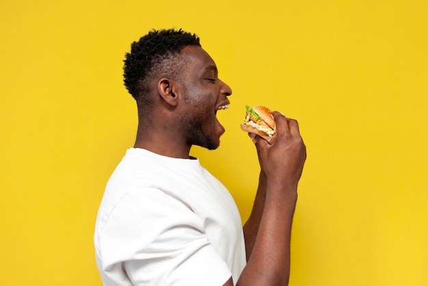 Un Afro-Américain en T-shirt blanc mord un gros hamburger sur un fond jaune isolé.