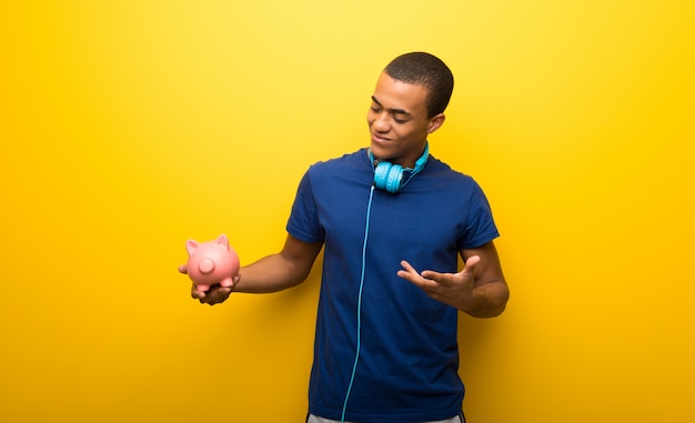 Afro-américain avec un t-shirt bleu sur un mur jaune tenant une grande tirelire