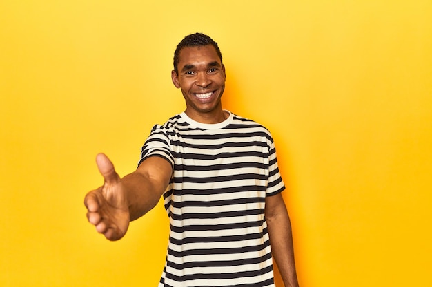 Un Afro-Américain en t-shirt rayé est un studio jaune tendant la main à la caméra dans un geste de salut.