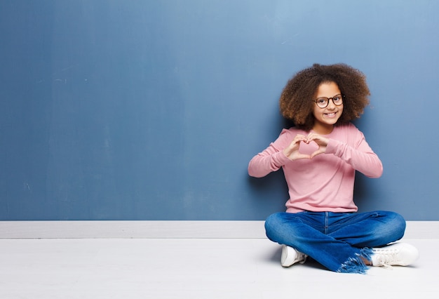 Afro-américaine petite fille souriante et se sentir heureuse, mignonne, romantique et amoureuse, en forme de cœur avec les deux mains assis sur le sol