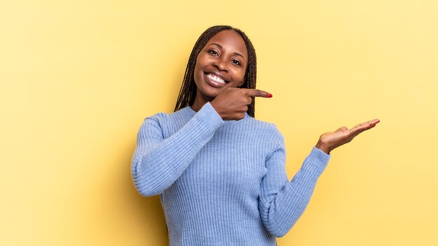 Afro black jolie femme souriante, se sentant heureuse, insouciante et satisfaite, pointant vers le concept ou l'idée sur l'espace de copie sur le côté