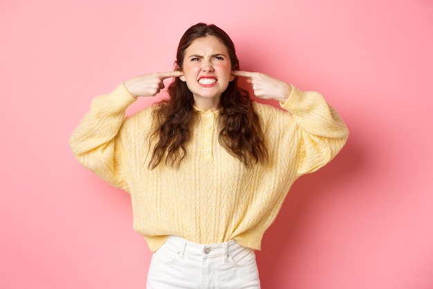 Agacé jeune femme maudissant les voisins en haut à la recherche des oreilles bouchées avec les doigts de bruit fort ennuyeux se plaindre de la musique debout contre le mur rose