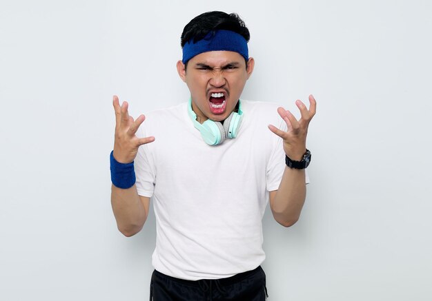 Agacé jeune sportif asiatique en bandeau bleu et t-shirt blanc avec un casque crie fort exprime la rage et la haine isolé sur fond blanc Concept de sport d'entraînement