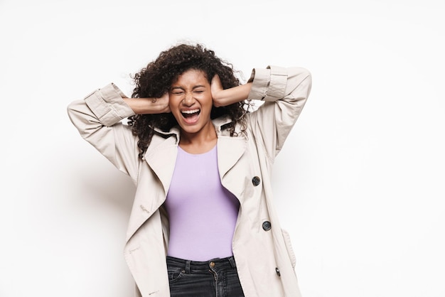 Agacé jolie jeune femme portant une veste d'automne debout sur un mur blanc, couvrant les oreilles, criant