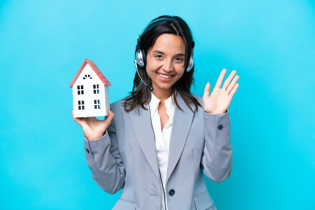 Agent immobilier hispanique tenant une maison de jouet isolée sur fond bleu saluant avec la main avec une expression heureuse
