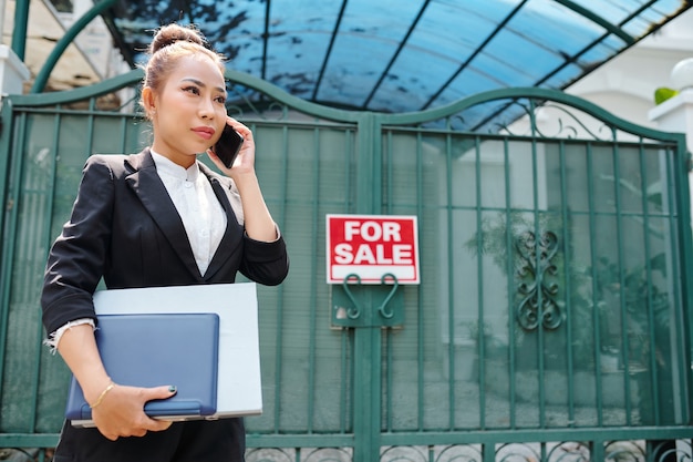 Agent immobilier vietnamien féminin avec des documents en mains debout aux portes de la maison en vente et parlant au téléphone