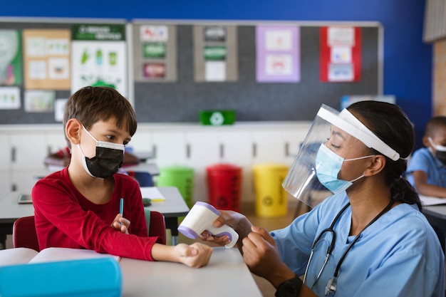Agent de santé féminin portant un écran facial mesurant la température d'un garçon à l'école primaire