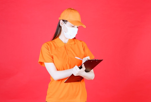 Agent de service féminin en code vestimentaire et masque de couleur orange tenant une feuille de projet et un stylo et prenant des notes