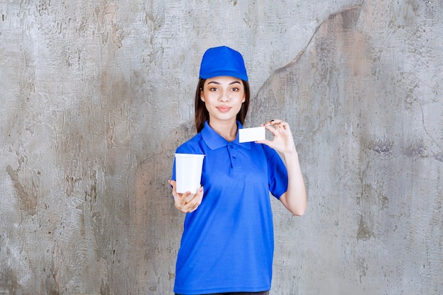 Agent de service féminin en uniforme bleu tenant un gobelet jetable blanc et présentant sa carte de visite.