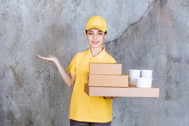 Agent de service féminin en uniforme jaune tenant un stock de boîtes en carton à emporter et de gobelets en plastique.