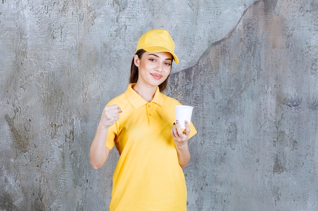 Agent de service féminin en uniforme jaune tenant une tasse en plastique et montrant un signe positif de la main.