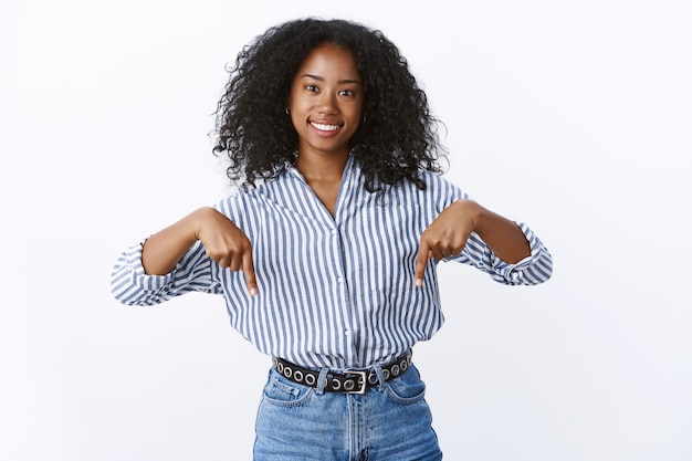 Agréable et agréable, belle jeune femme africaine aux cheveux bouclés de 25 ans portant un chemisier rayé pointant vers le bas souriant joyeusement vous demandant de vérifier, dépêchez-vous cliquez sur promo, mur blanc