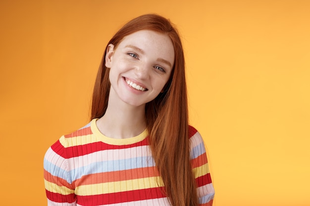 Agréable charismatique jeune fille charmante rousse insouciante confiante tête inclinable souriant largement dents blanches parler avec désinvolture bonne humeur exprimer des émotions heureuses positives debout fond orange.