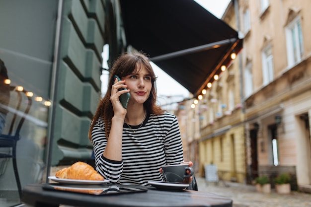 Agréable femme brune ayant une conversation mobile tout en se reposant sur la terrasse du café