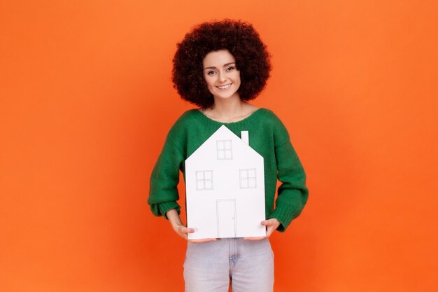 Agréable femme à la coiffure Afro portant un chandail vert de style décontracté tenant une maison en papier blanc, achetant un nouvel appartement, agence immobilière. Studio intérieur tourné isolé sur fond orange.