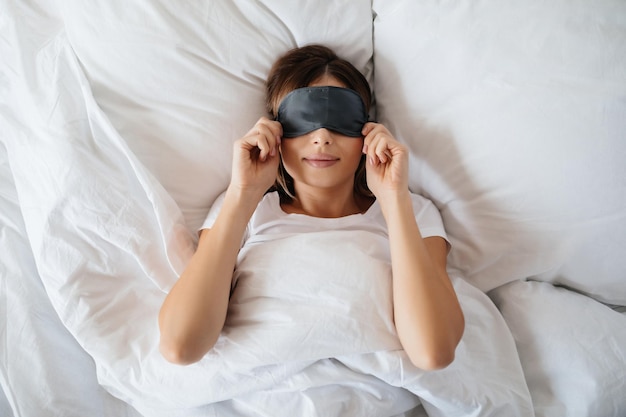 Agréable Jeune Femme Allongée Dans Son Lit Et Mettant Un Masque Pour Les Yeux Avant D'aller Dormir