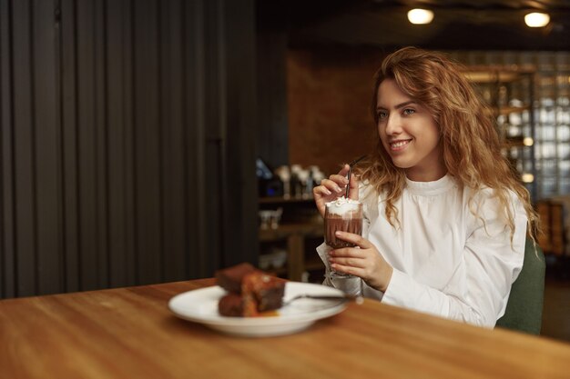 Agréable jeune femme aux cheveux blonds bouclés en chemisier blanc se détendre au café local, boire du cacao savoureux et dessert au chocolat sucré