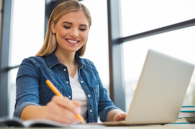 Agréable jeune femme intelligente assise devant l'ordinateur portable et étudie tout en se préparant pour un exemple