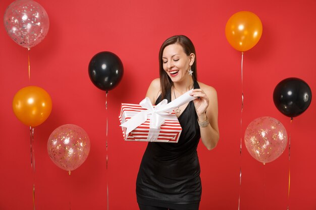 Agréable jeune femme en robe noire célébrant, ouvrant une boîte rouge avec un cadeau, présente sur des ballons à air de fond rouge vif. Saint-Valentin, bonne année, concept de fête de vacances de maquette d'anniversaire.