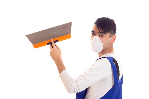 Agréable jeune homme aux cheveux noirs en chemise blanche et combinaison bleue avec respirateur blanc sur son visage à l'aide d'une spatule sur fond blanc en studio