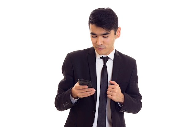 Agréable jeune homme aux cheveux noirs en chemise blanche et costume noir avec cravate à l'aide de son téléphone sur fond blanc