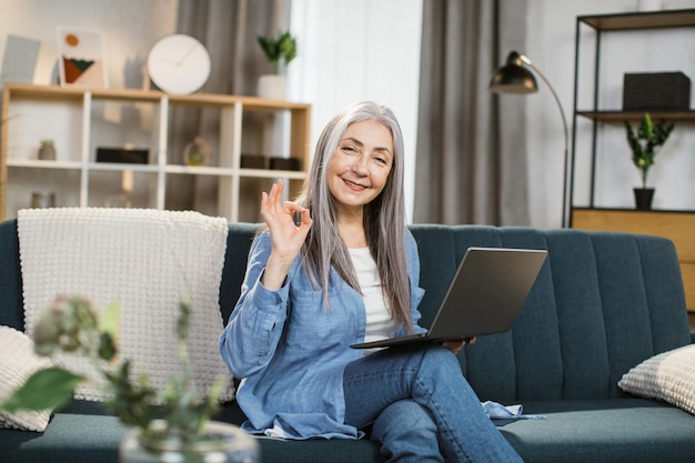 Agréable jolie femme aux cheveux gris aux cheveux longs assis sur un canapé avec un ordinateur portable sur les genoux