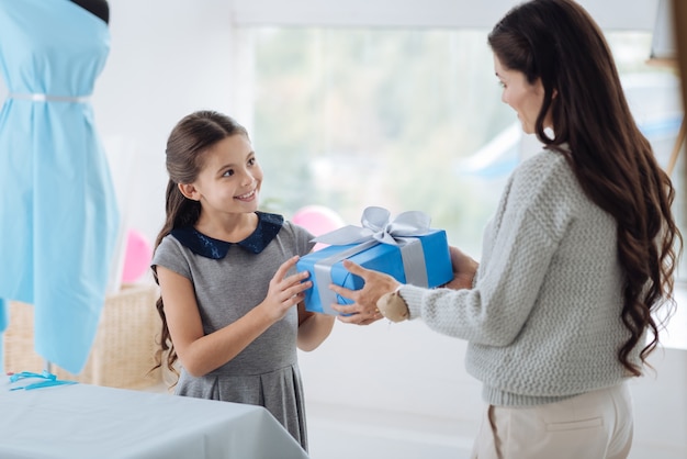 Agréable surprise. Bonne belle fille positive tenant une boîte-cadeau et souriant tout en recevant un cadeau d'anniversaire de sa maman