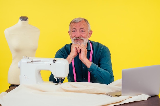 Agréable vieil homme travaillant dans un studio de couture faisant des vêtements à la machine à coudre, fond jaune.