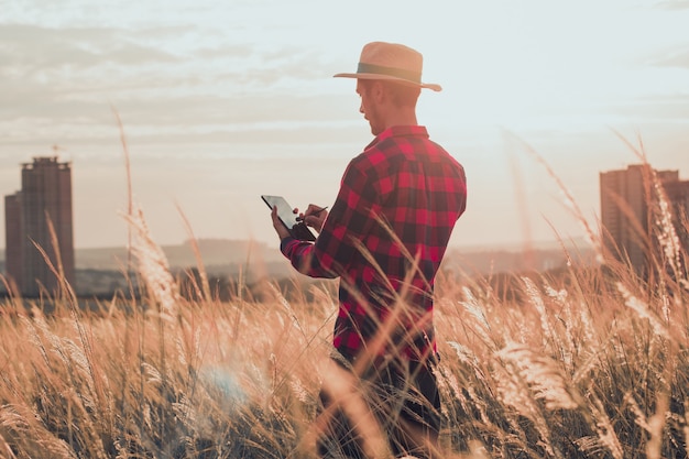 Agriculteur avec chapeau et tablette mobile analysant la plantation au coucher du soleil.