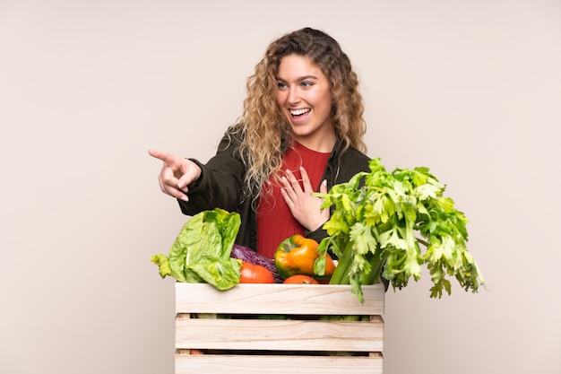 Agriculteur avec des légumes fraîchement cueillis dans une boîte isolée sur le mur de pointage du doigt beige sur le côté