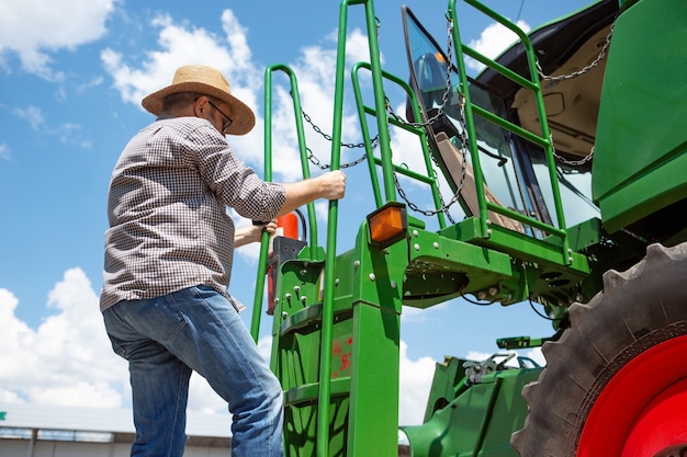 Agriculteur professionnel avec un tracteur moderne au processus de travail. Semble confiant, couleurs estivales vives, soleil. Agriculture, exposition, machines, production végétale. L'homme senior grimpe sur la machine.