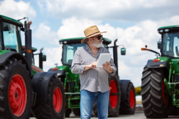 Agriculteur professionnel avec un tracteur moderne au travail avec tablette. Semble confiant, couleurs estivales vives, soleil. Agriculture, exposition, machines, production végétale. Homme supérieur près de sa machine.