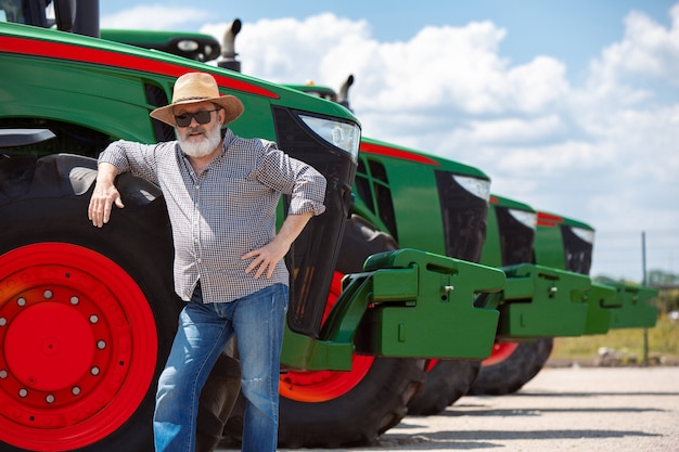 Agriculteur professionnel avec un tracteur moderne, combinez-vous dans un champ au soleil au travail. Couleurs d'été confiantes et lumineuses. Agriculture, exposition, machines, production végétale. Homme supérieur près de sa machine.