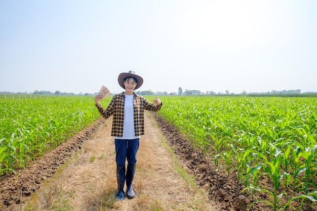 Une agricultrice asiatique dans une chemise rayée portant un chapeau posant debout avec des factures de revenu de maïs