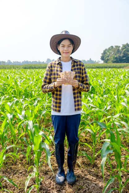 Une agricultrice asiatique porte une chemise debout et tient un billet de banque thaïlandais dans une ferme de maïs vert