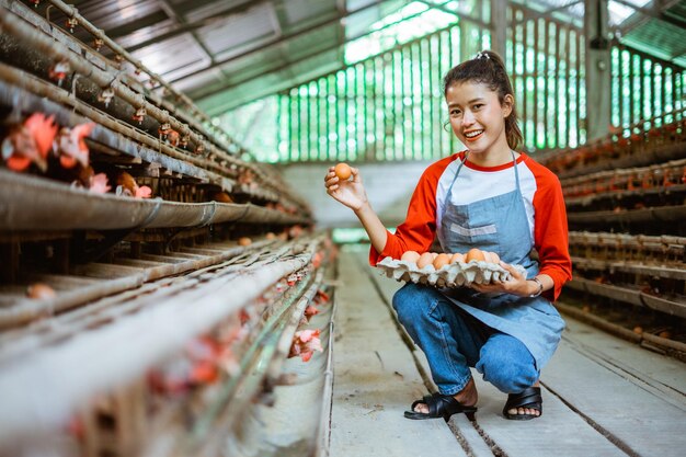 Une agricultrice asiatique souriante prend des œufs de la cage et porte un plateau accroupi près du