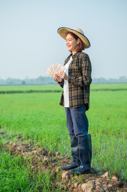 Une agricultrice asiatique vêtue de blouses brunes et de jeans se tient debout et pose en tenant des billets d'argent avec une expression souriante.