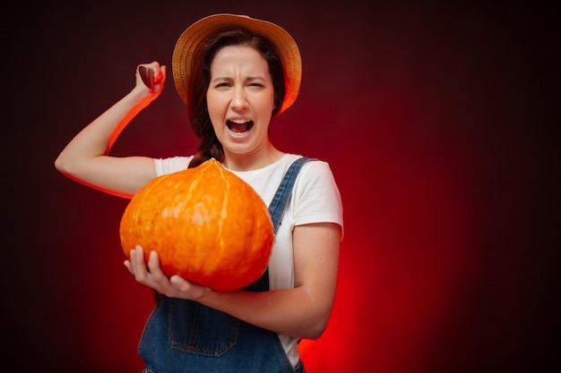 Une agricultrice attaque avec un couteau tranchant tenant une grande citrouille pour halloween sur fond rouge