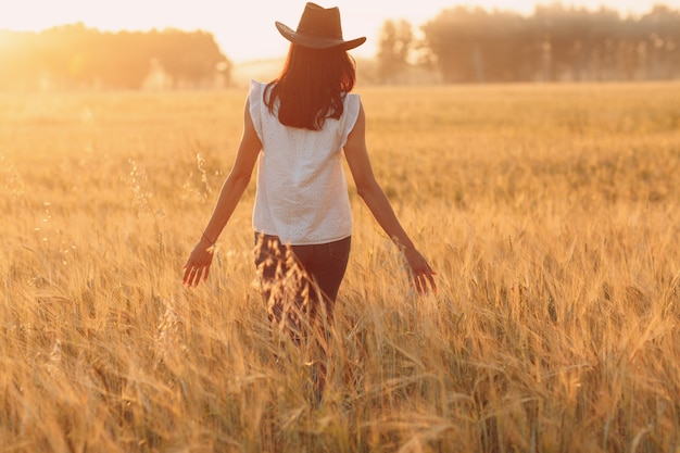 Agricultrice au chapeau de cowboy marchant avec les mains sur les oreilles au champ de blé agricole au coucher du soleil.