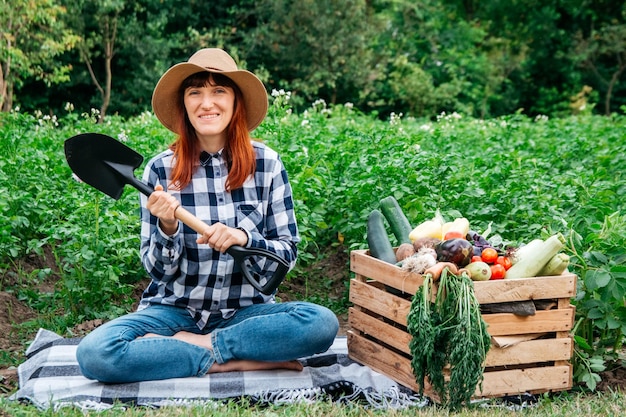 Agricultrice avec une pelle assise près de légumes biologiques frais dans une boîte en bois sur fond végétal