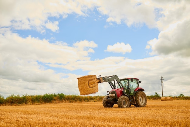Photo agriculture de paille et tracteur dans une ferme pour la durabilité sur un champ ouvert pendant la saison des récoltes de printemps nature ciel et nuages avec un véhicule agricole rouge récoltant du foin dans une campagne