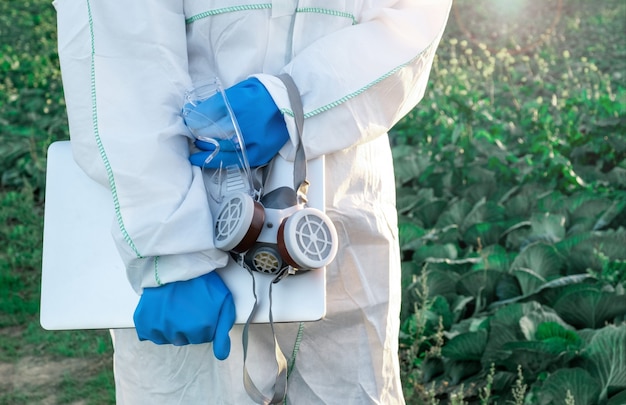 Un agronome vêtu d'une combinaison de protection blanche, d'un masque et de gants bleus tient un ordinateur portable contre le terrain