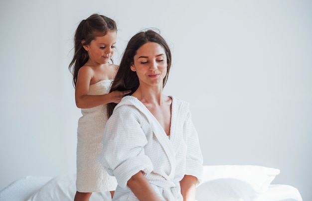 Aide à la coiffure. Une jeune mère avec sa fille a une journée de beauté à l'intérieur dans une salle blanche.