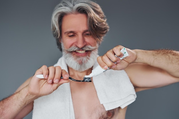 À l'aide de dentifrice Un homme âgé moderne et élégant aux cheveux gris et à la barbe est à l'intérieur