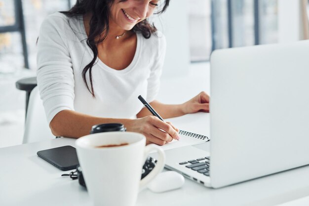 À l'aide d'un ordinateur portable et d'un bloc-notes Jeune femme indépendante travaillant à l'intérieur au bureau pendant la journée