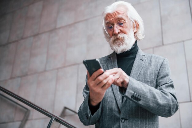 À l'aide d'un smartphone, un homme d'affaires senior en vêtements formels avec des cheveux gris et une barbe est à l'extérieur